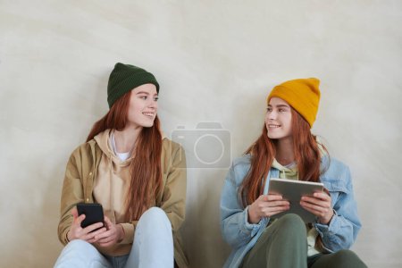 Photo for Studio portrait of two beautiful young sisters wearing casual clothes sitting together on floor surfing Interent on gadgets looking at each other - Royalty Free Image