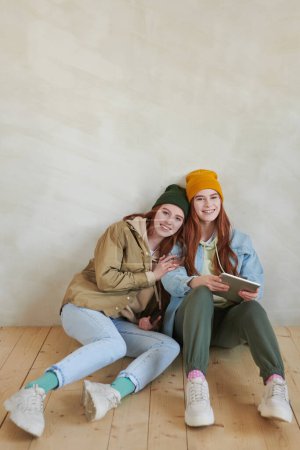 Photo for Vertical high angle studio portrait of joyful young female twins wearing stylish casual outfits sitting together on flloor smiling at camera - Royalty Free Image