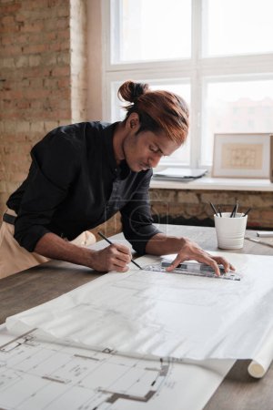Photo for Focused young mixed race engineer with ponytail leaning on desk and using ruler while drawing house plan in office - Royalty Free Image