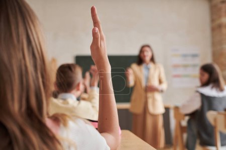 Téléchargez les photos : Gros plan d'une écolière méconnaissable levant la main en classe avec une enseignante en arrière-plan, espace de copie - en image libre de droit
