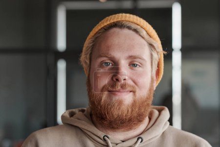 Headshot of satisfied young red-bearded Caucasian man in bright hat posing in modern office