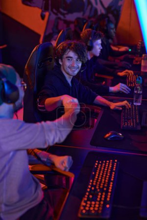 Photo for Satisfied young men sitting in esports bar with neon illumination and making fist bump while playing video game as team - Royalty Free Image