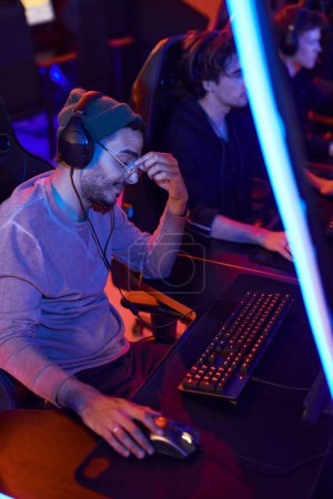 Photo for Tired mixed race esports gamer in headphones rubbing bridge of nose while playing online game in dark space - Royalty Free Image