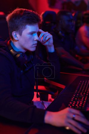 Photo for Concentrated Caucasian teenager with headphones around neck sitting at table in computer club and using modern computer - Royalty Free Image