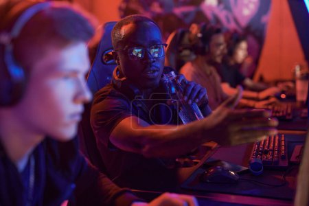 Photo for Skilled young African-American esports gamer in glasses gesturing hand and commenting action of guy in computer club - Royalty Free Image