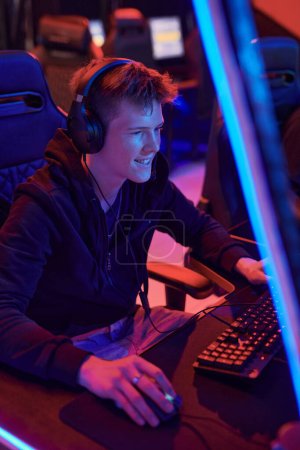 Photo for Excited young Caucasian gamer in headphones using powerful computer in esports club with neon illumination - Royalty Free Image