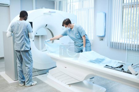 Téléchargez les photos : Infirmières multiethniques en masques et gommages préparant une IRM pour l'examen du patient : femme étalant une feuille jetable sur la table - en image libre de droit