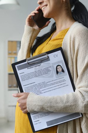Vertical image of young pregnant businesswoman with resume talking to new candidate on mobile phone