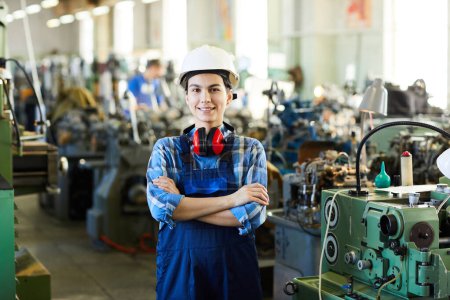 Foto de Feliz joven trabajadora atractiva en general de pie contra las máquinas industriales y sonriendo a la cámara - Imagen libre de derechos