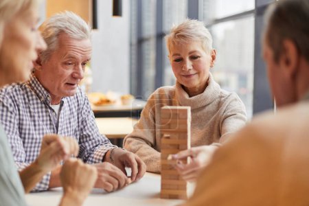 Foto de Grupo de contenido concentrado personas mayores sentadas en la mesa en el hogar de ancianos y jugando jenga juntos - Imagen libre de derechos
