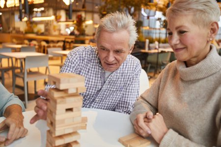 Group of concentrated senior friends sitting at table in nursing home leisure room and playing jenga