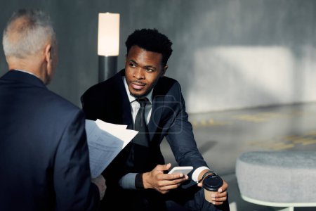 Foto de Joven empresario afroamericano confiado con teléfono inteligente y taza de café para llevar sentado en la sala y discutiendo datos con su colega - Imagen libre de derechos