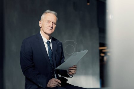 Foto de Retrato de ejecutivo senior estricto serio con el pelo gris sentado en el vestíbulo oscuro y la celebración de papeles de negocios - Imagen libre de derechos