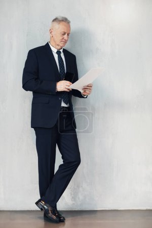 Foto de Pensativo experimentado hombre de negocios maduro en traje formal oscuro apoyado en la pared y analizando los puntos de contrato - Imagen libre de derechos
