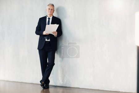 Foto de Retrato de un empresario sénior serio y exitoso en traje negro apoyado en la pared en el vestíbulo y leyendo periódicos - Imagen libre de derechos