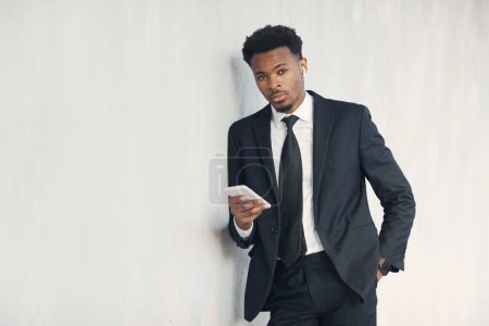 Foto de Retrato de un joven hombre de negocios negro guapo serio con barba apoyada en la pared y el uso de un teléfono inteligente para escuchar el mensaje de audio en auriculares - Imagen libre de derechos