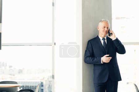 Foto de Serious pensativo confiado hombre de negocios senior en traje de pie por columna y discutir los problemas de trabajo por teléfono - Imagen libre de derechos