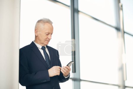 Foto de Serious ocupado empresario de pelo gris en traje oscuro apoyado en la pared en el vestíbulo moderno con ventanas panorámicas y tomar notas en el teléfono inteligente - Imagen libre de derechos