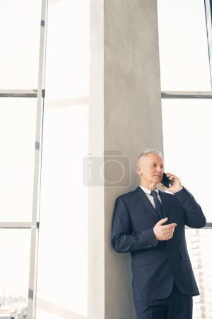 Foto de Contenido guapo hombre de negocios maduro en traje formal apoyado en la columna en el vestíbulo mientras habla por teléfono celular - Imagen libre de derechos
