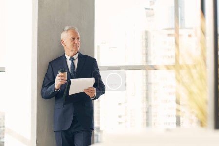 Foto de Pensativo hombre de negocios de pelo gris senior seguro en traje elegante examinar papeles y beber café en el vestíbulo de la oficina - Imagen libre de derechos
