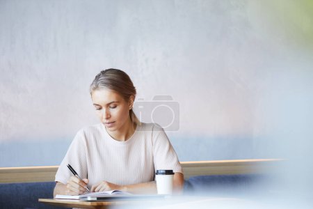 Téléchargez les photos : Femme blonde concentrée sérieuse en pull décontracté assis à la table et prenant des notes dans le journal tout en travaillant dans un café - en image libre de droit