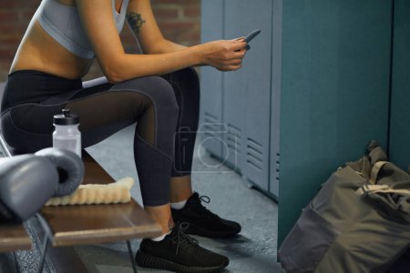 Photo for Unrecognizable woman in sportswear sitting on bench in gym dressing room using her smartphone - Royalty Free Image
