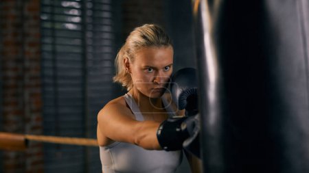 Téléchargez les photos : Portrait horizontal d'une femme forte frappant un sac lourd pendant un entraînement de boxe - en image libre de droit