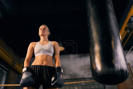 Téléchargez les photos : Plan horizontal à angle bas de la jeune femme blonde debout sur l'anneau avec un sac de boxe à côté d'elle - en image libre de droit