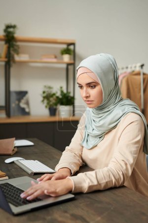 Photo for Vertical portrait of beautiful young woman wearing light blue hijab sitting at table working on laptop at home - Royalty Free Image