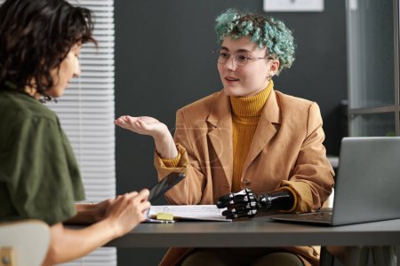 Foto de Joven mujer de negocios con brazo protésico hablando con el cliente durante su reunión en la oficina - Imagen libre de derechos