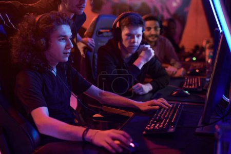 Photo for Group of concentrated team members looking at computer monitor and assisting guy to win game in neon-illuminated room - Royalty Free Image