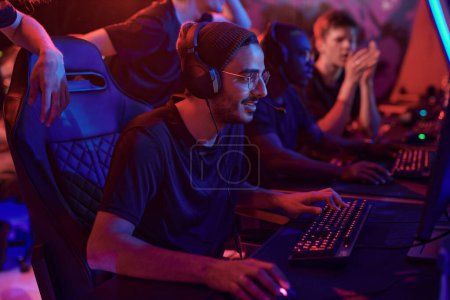 Photo for Smiling young professional gamer in hat and eyeglasses using computer while participating in esports tournament - Royalty Free Image