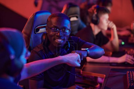 Téléchargez les photos : Souriant jeune homme noir dans des lunettes assis dans une chaise d'ordinateur et donnant coup de poing à un membre de l'équipe pendant qu'ils jouent ensemble à un jeu vidéo - en image libre de droit