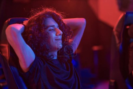 Photo for Gamer with long curly hair keeping eyes closed and holding hands behind head while relaxing in computer chair in esports bar - Royalty Free Image
