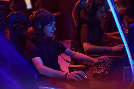 Photo for Young curly-haired gamer in hat and headset with microphone interacting with team in online game while using computer - Royalty Free Image
