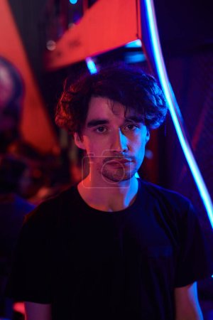 Photo for Portrait of sad young Caucasian man in black tshirt standing in esports bar, blue and red lights - Royalty Free Image