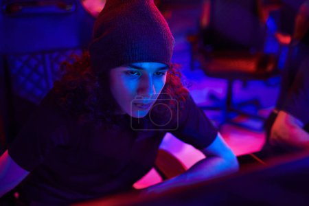 Téléchargez les photos : Concentré jeune développeur d'applications ou pirate en utilisant l'ordinateur dans la chambre bleu foncé - en image libre de droit
