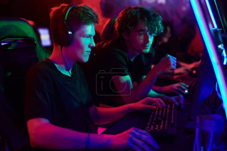 Téléchargez les photos : Curieux jeune homme dans les écouteurs jouer à un jeu d'ordinateur en ligne sous le contrôle de l'entraîneur cybersport dans le club de jeu - en image libre de droit