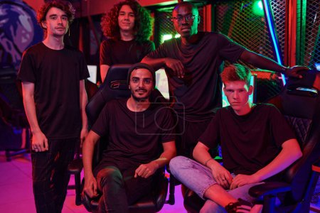 Téléchargez les photos : Equipe multiraciale de jeunes joueurs en t-shirts noirs posant ensemble dans un club de jeu, illumination rouge - en image libre de droit