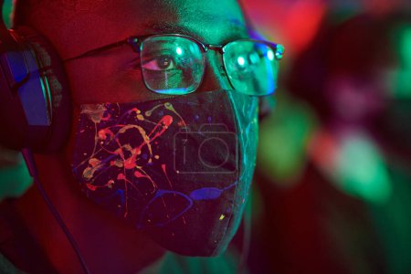 Téléchargez les photos : Jeune homme afro-américain sérieux dans des lunettes et masque facial élégant pensant misson jeu dans la lumière rouge - en image libre de droit