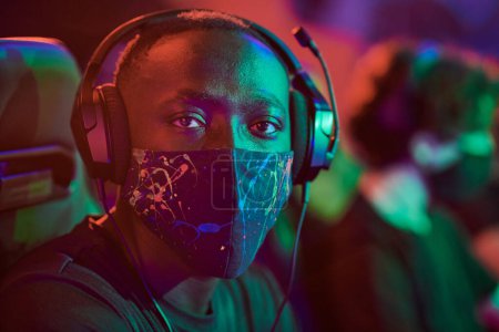 Photo for Portrait of young African-American esports gamer in headphones and abstract mask in red light - Royalty Free Image