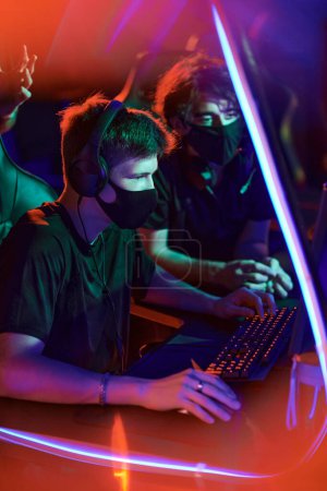 Photo for Concentrated young Caucasian man sitting at table in red light and using computer while participating in cybersport tournament - Royalty Free Image