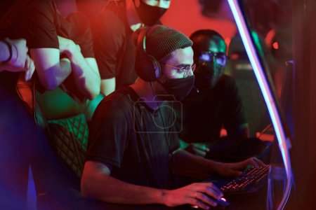 Téléchargez les photos : Concentré jeune Arabe gamer dans chapeau et masque en tissu jouant jeu vidéo tandis que les membres de son équipe le regardent - en image libre de droit