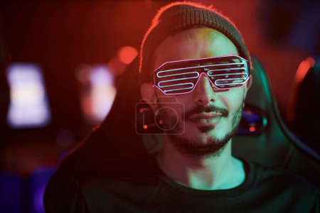 Téléchargez les photos : Portrait de contenu cool jeune homme métis en lunettes LED et chapeau assis dans le club informatique sombre - en image libre de droit