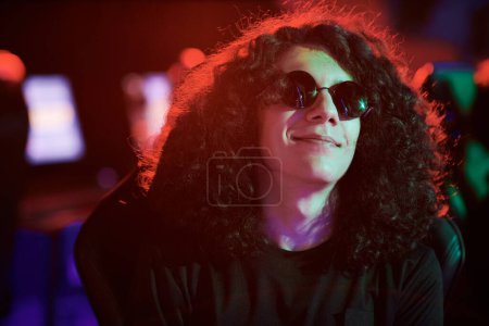 Photo for Smiling relaxed fashionable young Caucasian guy in round-shaped sunglasses sitting in red light in casino - Royalty Free Image