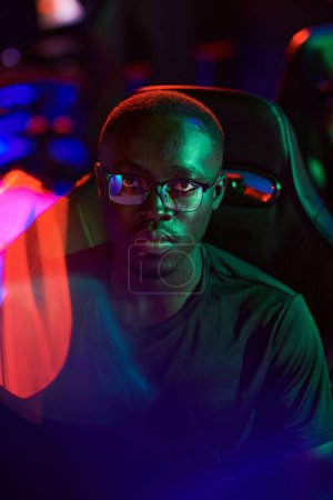 Téléchargez les photos : Portrait de jeune cybersportif noir sérieux participant dans des lunettes assis dans une chaise d'ordinateur dans des néons - en image libre de droit