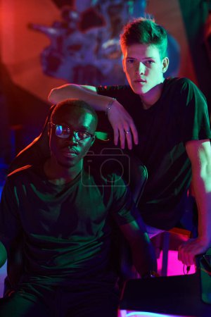 Photo for Young man leaning on computer chair with his African-American friend in modern computer club in neon light - Royalty Free Image