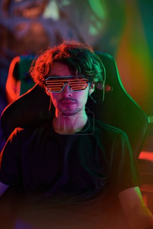 Photo for Portrait of serious handsome young gamer in glowing glasses sitting in modern armchair in room with neon illumination - Royalty Free Image