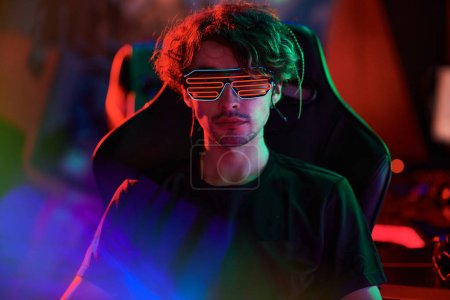 Téléchargez les photos : Portrait de sérieux jeune homme caucasien en néon rave lunettes assis dans la chaise d'ordinateur - en image libre de droit