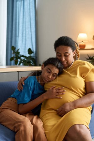 Téléchargez les photos : Adolescente embrassant sa mère enceinte alors qu'ils sont assis sur le canapé dans la chambre - en image libre de droit